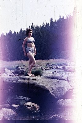 A woman in a patterned swimsuit stands on a rocky outcrop overlooking a forested landscape. Possibly a promotional or persona...