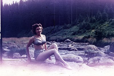 A young woman in a patterned bikini sits on rocks beside a rushing river. Pine trees form a backdrop, suggesting a mountain o...