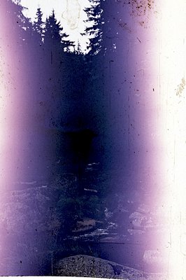 A faded, vertically-oriented photograph depicting a layered landscape. A stone structure or bridge is visible in the lower-ce...