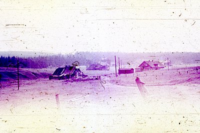 A snow-covered rural scene shows several modest houses and buildings against a backdrop of dense evergreen trees. Likely a sm...