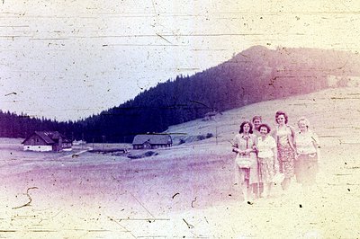 Four young women stand on a grassy slope, facing the camera. Behind them, a rural landscape features scattered homes and dens...