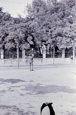 A grayscale photo shows a young man in shorts and a short-sleeved shirt observing a boy engaged in an activity beyond a low f...