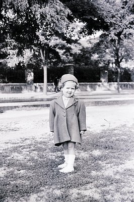 A young girl, circa 1960s, stands on a grassy area with a stone wall and trees in the background. She wears a double-breasted...