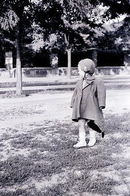 A young child, viewed from behind, stands on a grassy area. Wearing a warm coat, a distinctive hat, and sturdy boots, the chi...