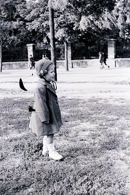 A young girl in a wool coat and hat stands on a grassy area, facing left. A park backdrop features stone pillars and distant ...