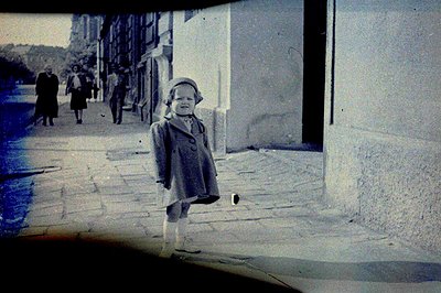 A young child in a coat and hat stands in a cobbled courtyard, facing the camera. Background shows figures and buildings. The...