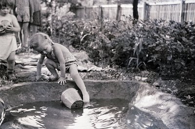 Young girl in a swimsuit kneels at the edge of a small, shallow pool, reaching for a bucket partially submerged in the water....