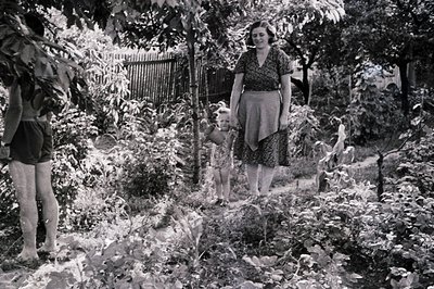 A mother and child stroll through a lush, overgrown garden. The woman wears a patterned dress and high-waisted skirt, indicat...