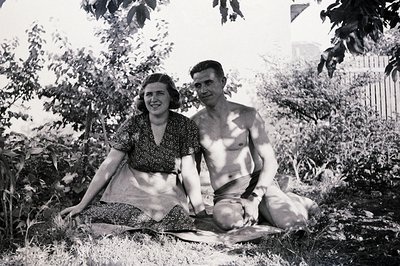 A couple sits on a blanket amid dense shrubbery, possibly a garden or seaside location. The woman wears a patterned dress; th...
