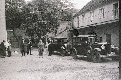 Formal portrait featuring a vintage Ford automobile & group of formally dressed individuals outside an apartment building. Ap...