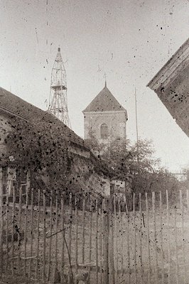 Rustic, weathered wood fencing obscures the view of a building and a tower, possibly a church or public structure. The archit...
