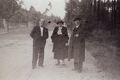 Three formally dressed figures stand on a dirt road bordered by tall trees. The individuals are posed in a seemingly candid m...