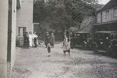 A group gathers outside a modernist building, circa 1930s. Two children play on a gravel drive flanked by vintage automobiles...