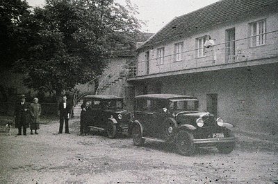 A 1930s black car sits prominently before a two-story building with a balcony. Four figures – two men in suits, two in more c...