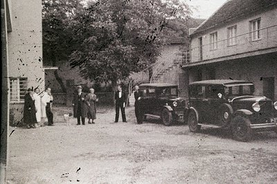 Group portrait featuring a vintage sedan and formally dressed individuals in a courtyard setting. The architecture suggests a...