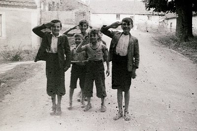 Four boys stand saluting on a dusty, rural road. The scene depicts simple clothing styles – one boy in a jacket, others in sh...