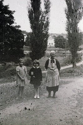 Three figures stand on a gravel path lined with tall trees: two children in vintage clothing, and an older woman in an apron....