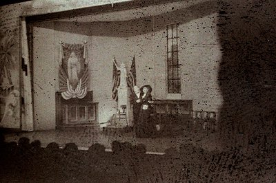 A partially obscured view of a formal interior, likely a government or civic building. Visible are draped flags, a large port...