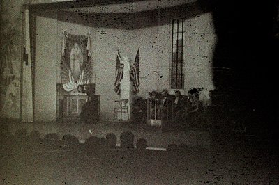 Formal interior scene: group assembled facing a raised platform with draped flags & what appears to be a religious icon. Like...