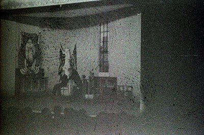 A grainy, grayscale image depicts an interior space with a large window and an ornate, possibly religious, hanging textile. S...