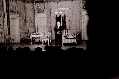 A stage scene with a man in a suit stands near a window draped with fabric. Furniture, including a settee and table, are arra...