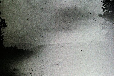 Monochrome landscape photo featuring a low hill or ridge, partially obscured by atmospheric haze or fog. Heavily aged; charac...