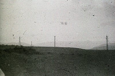 A panoramic landscape shows a grassy hillside leading to a hazy horizon with visible mountains. Several utility poles punctua...