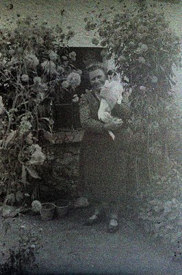 A young girl in a long dress stands in a courtyard, partially obscured by climbing vines and foliage. She appears to be holdi...