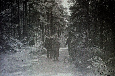 Three figures in long coats walk along a path through a dense forest. Appears to be a rural setting, possibly early 20th cent...