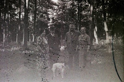 Formal portrait of a family—woman, man in a hat, and another man—accompanied by a dog in a wooded area. Likely a studio outdo...