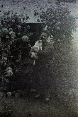 A grainy, black-and-white image captures a woman standing in a garden, obscured by dense foliage. She appears to be tending t...