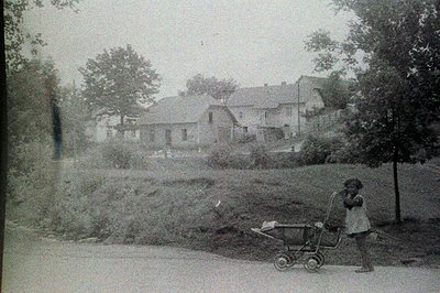 A young child stands near a wheeled toy, facing a large, multi-story building with numerous windows. Sloping ground and folia...