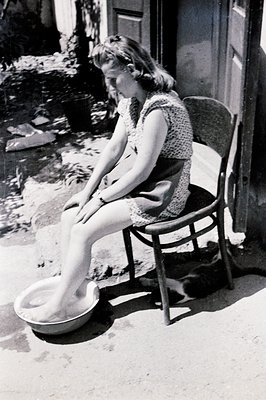 A woman sits on a wooden chair, soaking her feet in a round basin outside a building, likely a domestic setting. The scene su...