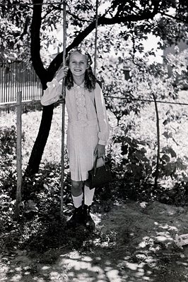 Young girl in a pleated dress and Mary Jane shoes stands in a garden. She holds a small bag, hair adorned with a ribbon. Like...