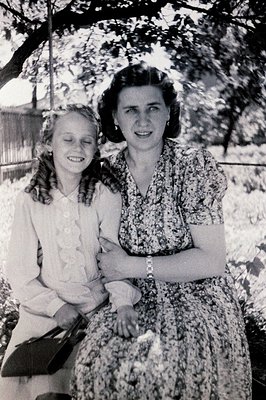 A young girl and woman, likely mother and daughter, pose outdoors. The woman wears a floral print dress and a wristwatch; the...