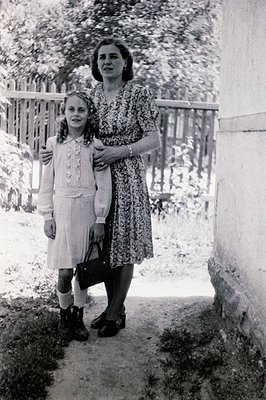 A young girl and woman stand near a weathered stone wall and wooden fence. The woman wears a patterned, mid-length dress and ...