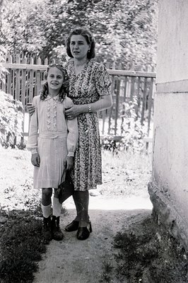 A young girl and woman stand close together in a garden setting. The girl wears a dress and boots, while the woman’s floral p...