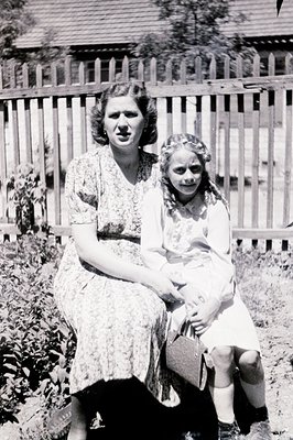 A mother and young daughter pose for a formal portrait, seated before a rustic wooden fence. The woman wears a patterned flor...