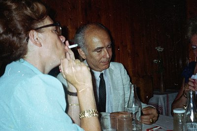 A man in a suit and tie smiles across a table from a woman in a light blue shirt, who is smoking a cigarette. Vintage color p...