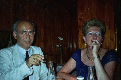 A man and woman sit at a table in a wood-paneled room. The man, in a light suit and tie, holds a cigarette; the woman wears a...