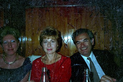 Three individuals seated at a table with wooden paneling. Woman in black, center in a red dress, man in a suit and tie. Likel...