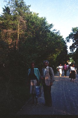 Formal portrait on a rural road, featuring an older couple in 1970s attire—she in a patterned skirt and teal jacket, he in a ...