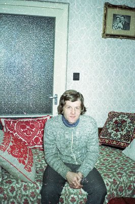 A man sits cross-legged on a patterned sofa within a room exhibiting 1970s décor. He wears a fair isle sweater and sits befor...