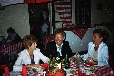 A warmly lit scene captures three individuals dining at a table covered with a red and white checkered cloth. The man in a su...