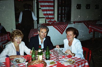 Three individuals seated at a restaurant table covered with a red and white checkered tablecloth. Plates of food, bread, and ...