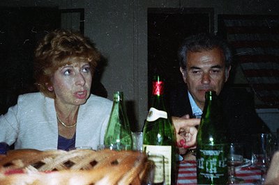 A candid, slightly grainy color photo shows a middle-aged couple at a table setting with mineral water bottles. The woman dis...