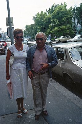 A smartly dressed couple poses beside a vintage car, likely in the 1970s. The woman wears a white dress & sandals, carrying a...