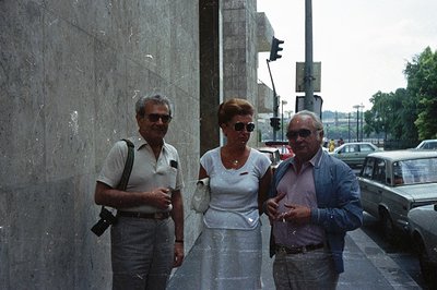 Three individuals, two men and a woman, stand against a monumental concrete wall. The men wear sunglasses and the woman carri...