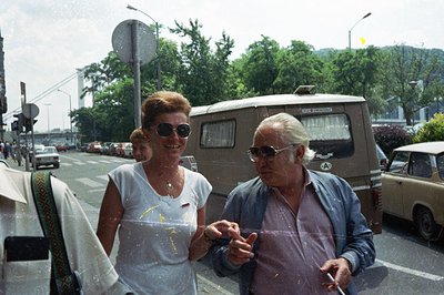 A candid street scene captures a smiling woman and a man with glasses. He smokes; a vintage van and car are visible alongside...