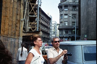 A couple strolls down a European street; the woman in a white top, the man photographing with a vintage camera. Background sh...